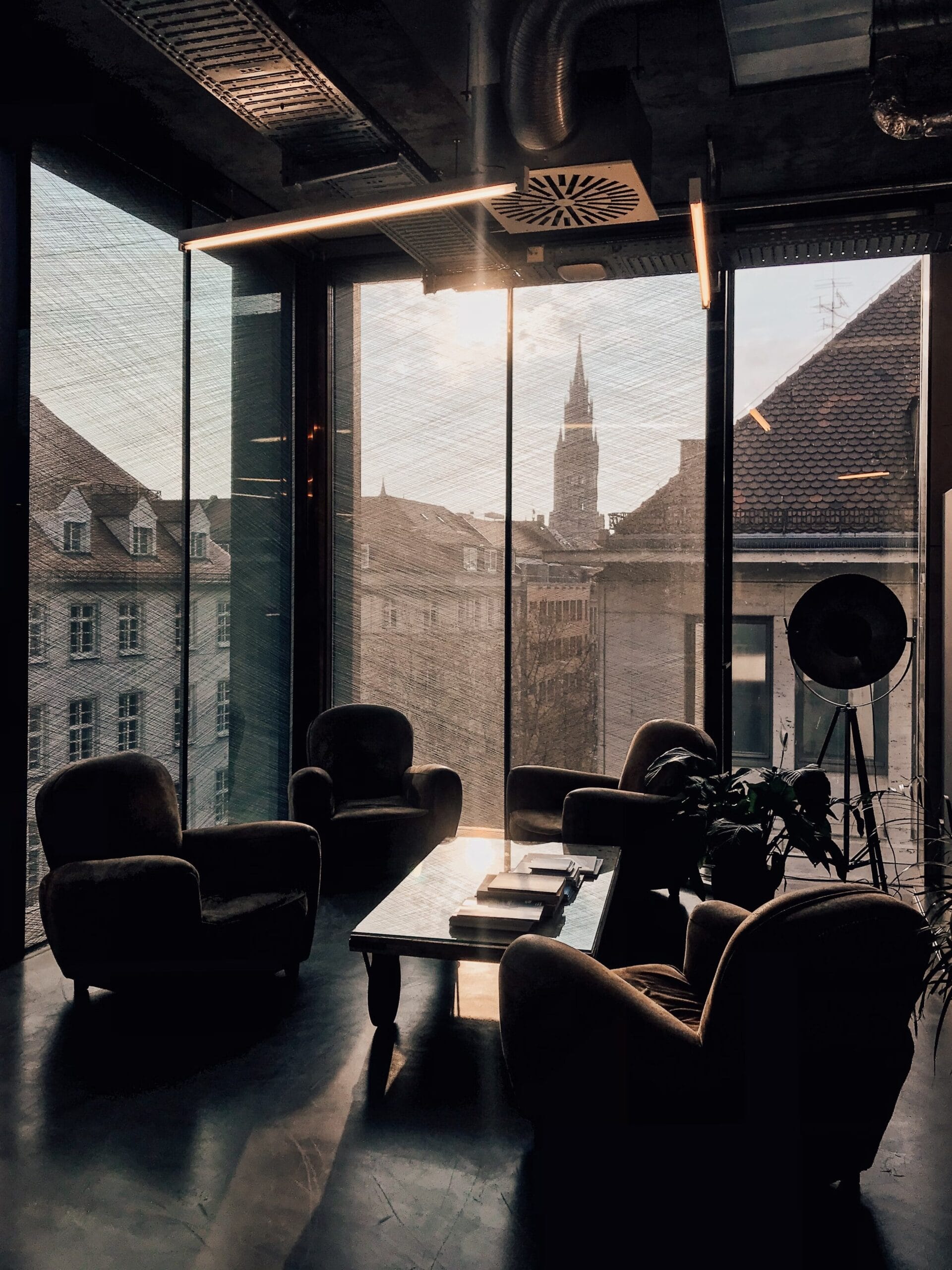 Vue intérieure d’un salon moderne avec des fauteuils confortables disposés autour d’une table basse. De grandes fenêtres du sol au plafond offrent une vue panoramique sur une ville européenne avec des bâtiments historiques et une flèche d’église en arrière-plan. La pièce présente des détails industriels au plafond et un projecteur au design saisissant.