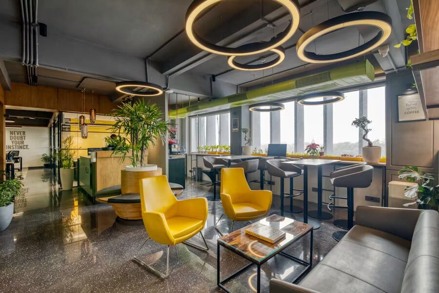 Espace de coworking avec luminaires circulaires suspendus, fauteuils jaunes, comptoirs avec tabourets et nombreuses plantes vertes.